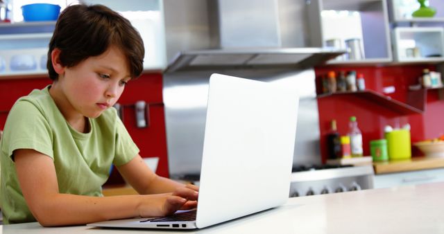 Young boy sitting at kitchen counter using laptop with focused expression in modern kitchen. Perfect for educational content, online learning platforms, technology tutorials, or advertisements featuring children and technology.