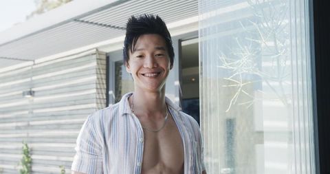 Smiling asian man standing in modern glass entry wearing open striped shirt and silver chain