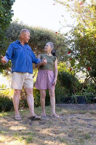Asian senior couple holding hands while walking on patchy lawn in sunlit backyard