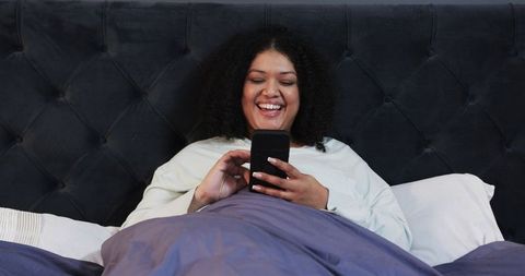 Relaxed African American Woman Laughing While Using Smartphone Under Duvet in Cozy Bedroom