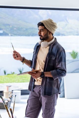 Young artist painting at waterfront studio wearing beanie and headphones holding palette