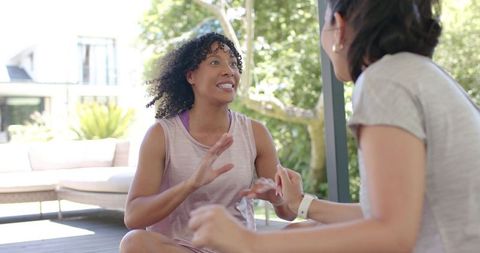 Women chatting after workout on sunlit patio, casual fitness conversation with smartwatch