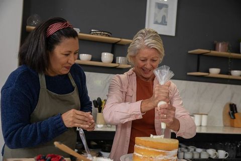 Multigenerational friends piping buttercream on three-layer sponge cake in cozy kitchen