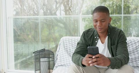 African american man using smartphone while sitting on sofa by window in natural light