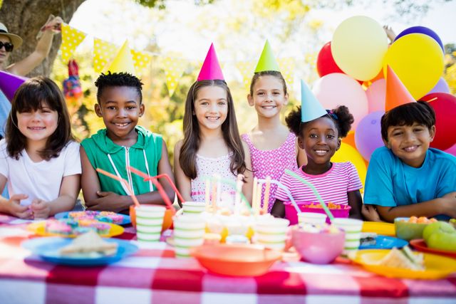 Group of children enjoying a birthday party outdoors, wearing colorful party hats and smiling. Ideal for use in advertisements, social media posts, and articles related to children's parties, celebrations, and outdoor activities. Perfect for promoting party supplies, children's events, and community gatherings.