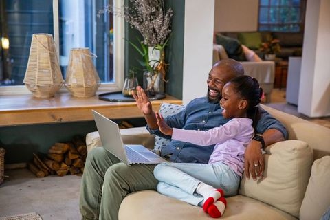 Father and daughter waving and laughing during video call on cozy living room couch