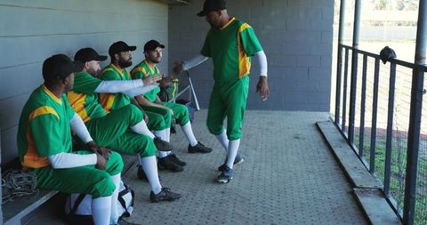 Baseball players sitting in dugout reaching toward walking teammate in green uniforms