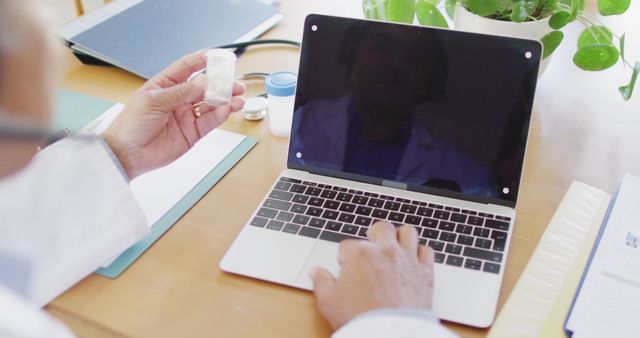 Doctor is holding a prescription bottle while using a laptop, suggesting a virtual medical consultation. This can be used to illustrate online healthcare, telemedicine, or remote consultations. It highlights how technology integrates into modern healthcare practices. Ideal for promoting digital health services, medical advice websites, and healthcare technology companies.