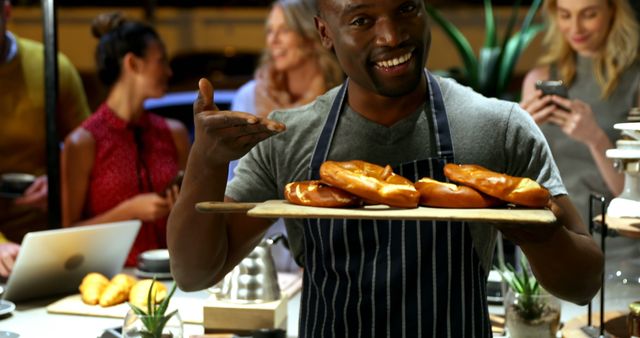A baker in an apron holding a tray of freshly baked pretzels and smiling while presenting them to customers in a modern bakery. Other customers are engaging in the background, creating a lively atmosphere. Ideal for use in marketing materials for bakeries, food promotions, culinary blogs, and food-related advertisements.