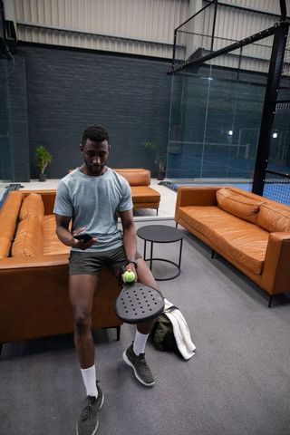 African american man using phone while holding padel paddle and ball in modern sports lounge