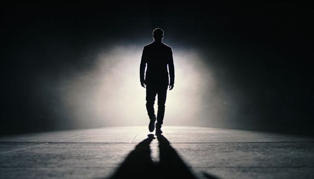 Silhouette man striding toward spotlight on foggy stage, dramatic backlit cinematic scene