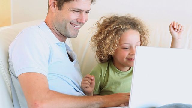 Father and young boy enjoy watching a video on a laptop in a relaxed home environment sitting on a couch. Ideal for concepts of family bonding, happy moments, father-son relationship, and the joy of parenting during technology use.