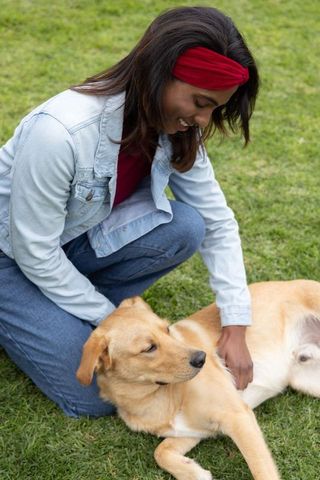 Indian woman kneeling on grass petting tan dog wearing denim jacket and red headband