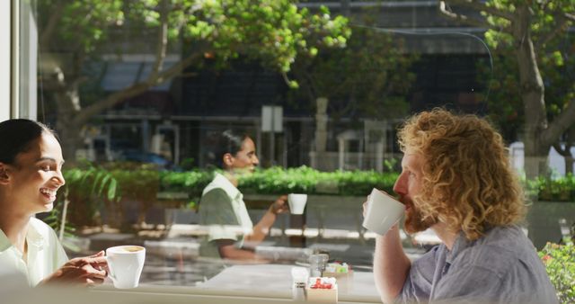 Interracial couple sitting at a sunny outdoor cafe drinking coffee and engaging in cheerful conversation. Ideal for themes including relationships, leisure, multicultural interaction, urban life, and relaxed moments in nature. Can be used in advertisements for cafes, social media content promoting inclusion, and lifestyle blogs focusing on everyday happiness.