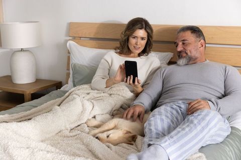 Middle-aged couple relaxing in bed sharing smartphone and petting puppy, cozy morning