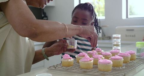 African grandmother and granddaughter decorating pink-frosted cupcakes in bright kitchen