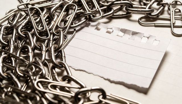 Interlocking steel chains surrounding torn lined paper with scattered paper clips