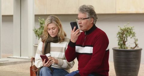 Middle-aged couple checking smartphones while waiting at urban transit stop bench