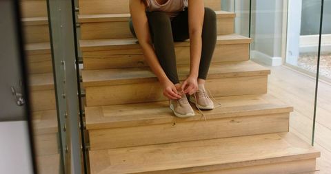 Woman tying sneaker laces on sunlit modern wooden staircase wearing olive leggings