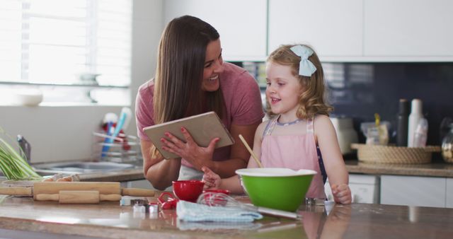 Mother and daughter are baking together in kitchen, using tablet for recipe. Highlights family bonding moments and interaction. Suitable for content related to cooking, family activities, digital recipes, and parenting.