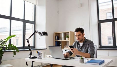 Typing man checking smartphone at bright minimalist home office desk with laptop and plants