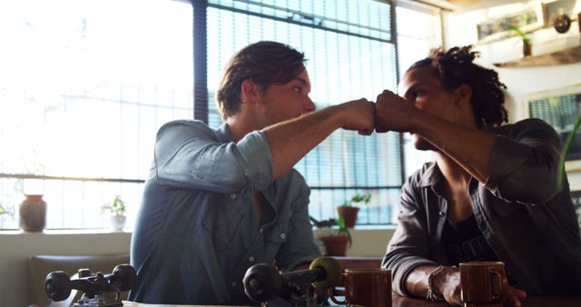 Two young men, one Caucasian and the other African American, are engaged in a friendly fist bump across a table, with copy space. They appear to be enjoying a casual meeting or celebration in a setting with natural light filtering through the window.