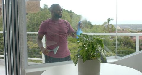 Man cleaning balcony glass door with spray bottle and squeegee near potted plant