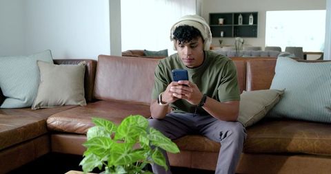 African american man listening with headphones while scrolling smartphone on leather sofa