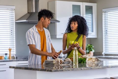 Unpacking reusable groceries in modern sunlit kitchen, diverse couple holding apples and oil