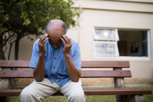 Elderly man sitting on a bench outdoors with his head in his hands, appearing upset and stressed. Suitable for use in articles or advertisements related to mental health, elderly care, loneliness, and retirement living.