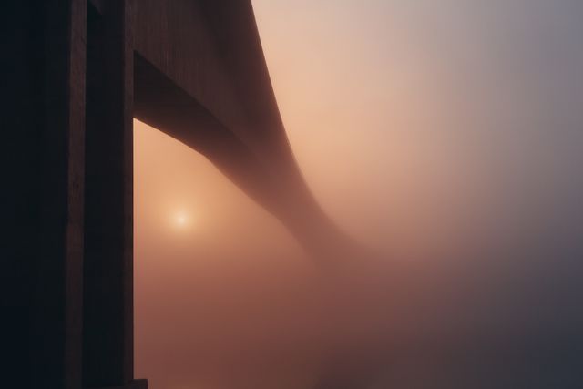 Concrete bridge structure seen disappearing into dense fog as the sun rises, creating a mysterious, serene atmosphere. Ideal for creating mood in architectural, travel, or nature-oriented projects. Suitable for blog posts, magazines, or advertisements focusing on early mornings, tranquil moments or evocative landscapes.