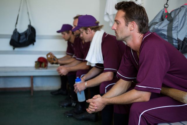 This image shows a group of tired baseball players sitting on a bench in a locker room. They are wearing maroon uniforms and appear to be taking a break, with one player holding a water bottle. This image can be used to depict themes of teamwork, sportsmanship, and the physical demands of athletic competition. It is suitable for articles, advertisements, and promotional materials related to sports, fitness, and team dynamics.