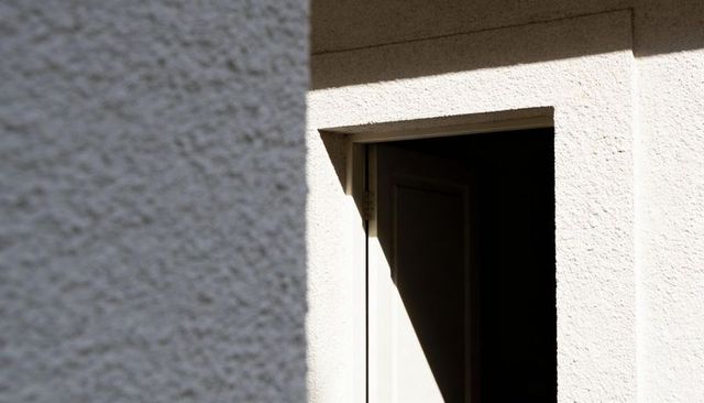 Ajar door casting triangular sun shadow on pebble stucco facade, minimalist architecture