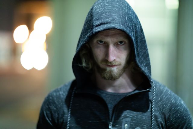 Fit Caucasian man with long blonde hair wearing sportswear and hoodie, exercising outdoors in the city at night. He is standing and looking straight at the camera with a determined expression. This image is ideal for use in fitness, sports, and urban lifestyle promotions, as well as advertisements for athletic wear and outdoor activities.