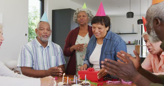 Senior friends are gathered together celebrating a birthday party, with one woman presenting a gift. The scene is festive with cupcakes, party hats, and cheerful expressions. Perfect for themes of friendship, community celebrations, aging, and social gatherings amongst diverse elderly individuals.