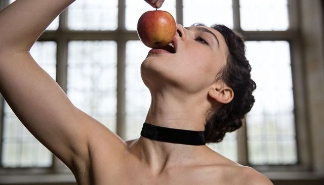 Woman tilting back biting red apple wearing black velvet choker in soft window light