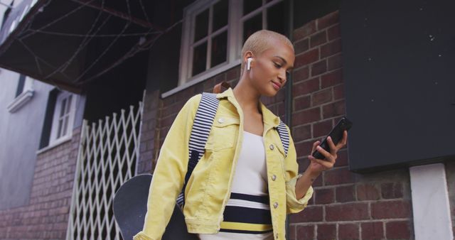 Urban youth walking down a city street, wearing wireless earbuds, holding skateboard, checking phone. Person dressed in yellow jacket and trendy attire reflects modern, independent lifestyle. Suitable for themes related to youth culture, technology, fashion, city living, and active lifestyle.