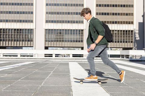Male skateboarder pushing on urban rooftop plaza holding smartphone with backpack, midday
