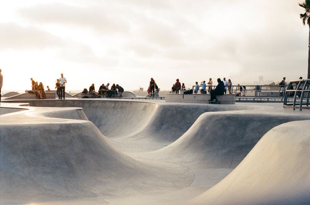 Group of skateboarders gathered at a concrete skatepark during sunset. Smooth ramps and curves of the park create an interesting texture in the foreground, with people watching and skating in the background. Ideal for use in advertisements for urban sports, youth clothing brands, or articles about skateboarding culture and outdoor activities.
