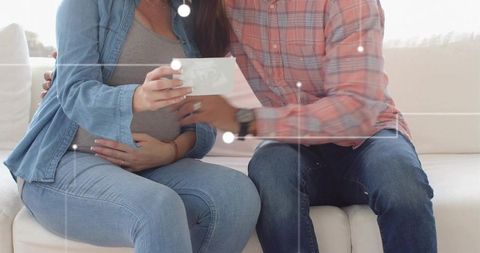 Expecting couple holding ultrasound print, pregnant woman cradling belly on light sofa