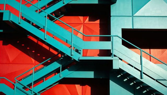 Teal Staircase Crossing Diagonal Shadows on Red Geometric Facade Industrial Architecture
