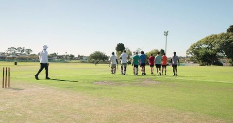 Cricket team walking across sunlit field with coach and pads on practice pitch
