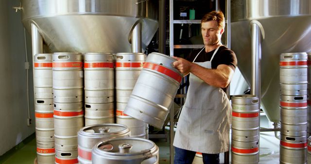 A middle-aged Caucasian man works in a brewery, handling a metal keg with care, with copy space. His profession as a brewer is evident from the stainless steel tanks and kegs surrounding him, indicating a craft beer production environment.