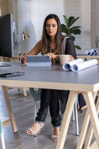 Indian woman working at modern studio desk using tablet with rolled blueprints