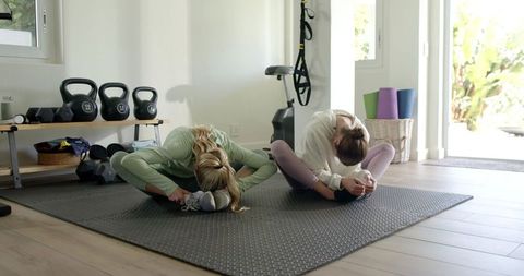 Two women doing butterfly forward fold stretch on home gym mat with kettlebells