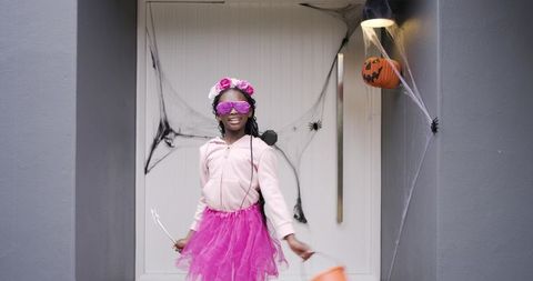 African american girl posing at doorstep in pink hoodie and tutu holding halloween pumpkin