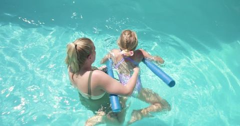 Parent teaching child to swim using foam pool noodles in sunlit turquoise backyard pool