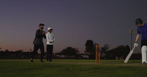 Evening cricket match at dusk showing batter adjusting bat at crease, bowler and wickets