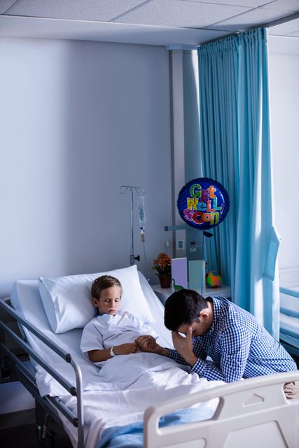 Father holding his son's hand beside hospital bed. Blue curtains and get well soon balloon in room. Used for topics on family, emotional support, healthcare, illness, recovery, and hospital visits.