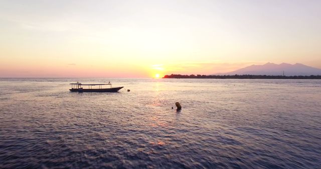 Person swimming near a boat during a calm sunset over the sea. Ideal for travel blogs, vacation destination promotions, relaxation and meditation content, and nature documentaries. The scene conveys peace and tranquility, perfect for illustrating leisurely or idyllic scenarios.